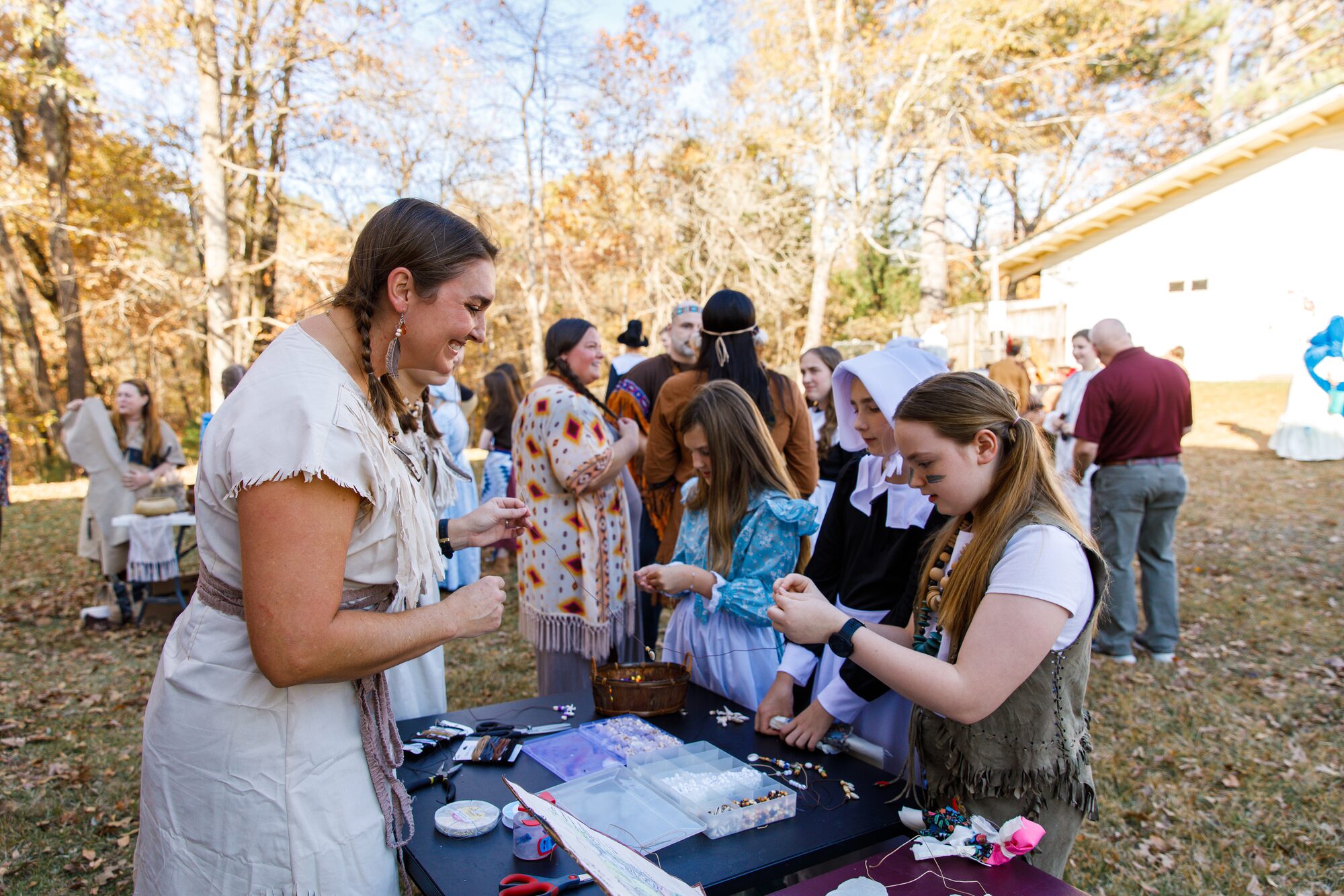 Students in colonial and Native American costumes making beaded crafts outdoors